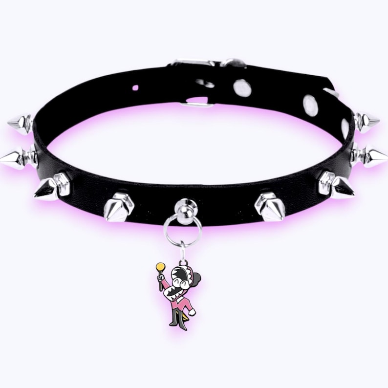 May include: A black collar featuring a series of silver-toned metal spikes arranged around the band. The collar includes a central silver ring holding a small, colorful enamel charm depicting a cartoon character in a pink suit holding a gold staff.