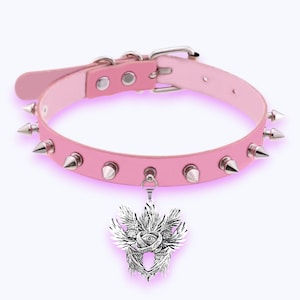 May include: Pale pink choker necklace with silver-tone spikes and a pendant. The pendant has a detailed design with wings and an eye. The choker has a buckle fastening and is made of a light pink material.