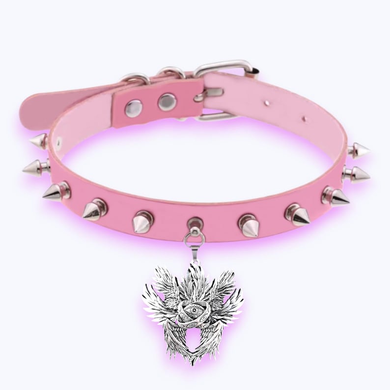 May include: Pale pink choker necklace with silver-tone spikes and a pendant. The pendant has a detailed design with wings and an eye. The choker has a buckle fastening and is made of a light pink material.