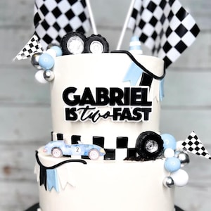 May include: A two-tiered white cake decorated with a checkered flag theme. The top tier has the words "Gabriel is two fast" in black lettering. The bottom tier has a checkered flag pattern and two blue race cars. The cake is surrounded by blue and silver balls, checkered flags, and small race car figurines.