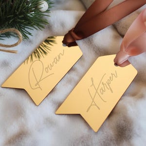 Könnte beinhalten: Zwei goldfarbene Geschenkanhänger mit den Namen "Rowan" und "Harper" in kursiver Schrift graviert. An jedem Anhänger ist ein braunes oder rosa Band befestigt. Die Anhänger liegen auf einer weichen, weißen Oberfläche, mit einem Tannenzweig und einem Tannenzapfen im Hintergrund.