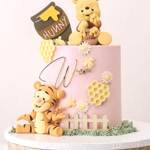 May include: A pink cake decorated with Winnie the Pooh and Tigger figurines, honeycombs, and daisies. The cake has the name "Wren" in gold script. The cake sits on a white-topped wooden cake stand.