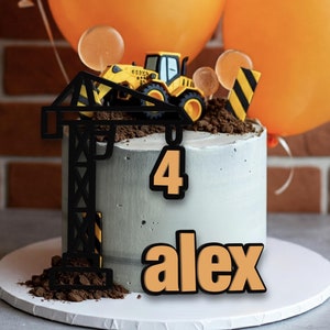 Op de afbeelding: Een grijze en witte taart versierd met een zwarte bouwkraan, een gele speelgoed-kipper en oranje ballonnen. De taart is versierd met het nummer "4" en de naam "alex" in oranje.