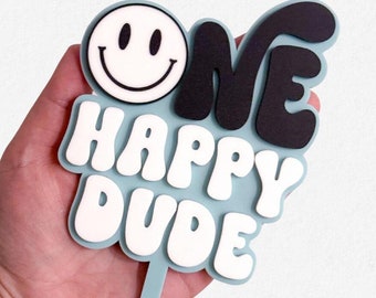 May include: A blue cake topper with the words "One Happy Dude" in white and black letters. A smiley face is on the left side of the topper.