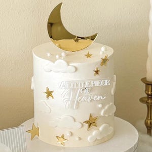 Pode incluir: Um bolo branco decorado com estrelas douradas, nuvens e uma lua crescente dourada. O bolo tem as palavras "A Little Piece of Heaven" escritas. Duas velas brancas em castiçais dourados ladeiam o bolo. Uma citação emoldurada e uma bandeja de biscoitos estão em primeiro plano.