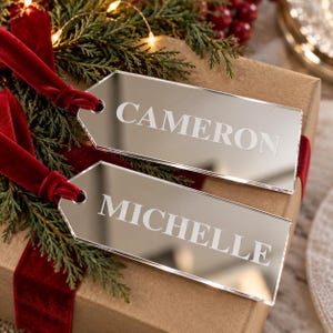 May include: Two rectangular mirror-finish acrylic gift tags with beveled edges and a hole at one end. Each tag is engraved with a name, CAMERON and MICHELLE, in white capital letters. A deep red velvet ribbon is looped through each tag hole.
