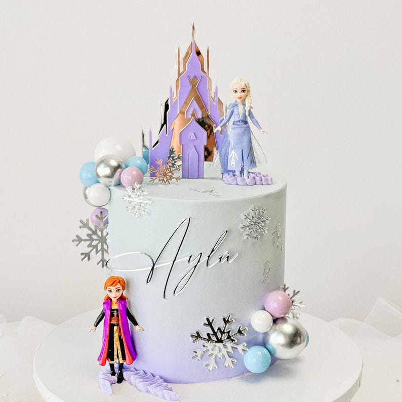 May include: A purple and blue ombre cake decorated with silver snowflakes, silver and blue balls, and two Frozen figurines. The cake is topped with a purple castle and the name "Ayla" is written in silver icing.
