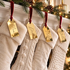 May include: Four cream-colored Christmas stockings with varied textures, including faux fur and cable knit patterns. Each stocking hangs from a red velvet ribbon attached to a gold-toned, rectangular metal name tag engraved with Mom, Dad, Violet, and Stella.