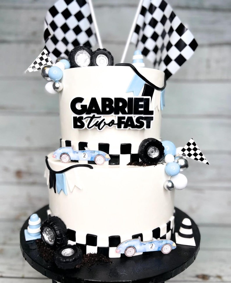 May include: A two-tiered white cake decorated with a checkered flag theme. The top tier has the words "Gabriel is two fast" in black lettering. The bottom tier has a checkered flag pattern and two blue race cars. The cake is surrounded by blue and silver balls, checkered flags, and small race car figurines.