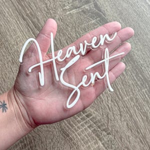 Pode incluir: Placa de acrílico transparente com as palavras "Heaven Sent" em fonte cursiva branca. A placa é segurada em uma mão contra uma superfície de madeira marrom clara. A placa é um item decorativo.