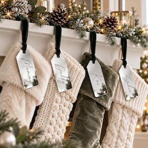 May include: Four Christmas stockings in cream faux fur, cream knit, olive velvet, and cream cable knit textures. Each stocking features a reflective silver acrylic tag hanging from a dark green velvet ribbon, inscribed with the names Mommy, Daddy, Kai, and Sadie.