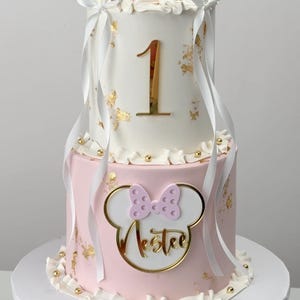 Puede incluir: Tarta de cumpleaños de dos pisos con glaseado blanco y rosa. El piso superior tiene un número 1 dorado, detalles de hojas doradas y un diseño de oreja de Minnie Mouse. El piso inferior presenta un diseño de Minnie Mouse con el nombre "Lester" en dorado.