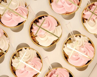 May include: A close-up of cupcakes decorated with pink and white frosting and gold crosses. The cupcakes are arranged in a box with white polka dots.