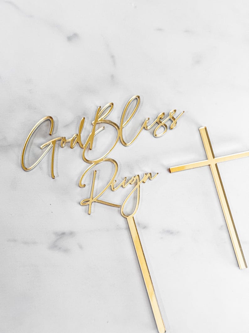 May include: Gold cake topper with the words "God Bless Reign" and a gold cross.