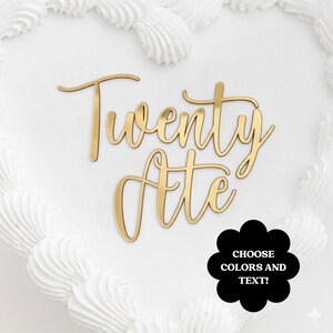 Op de afbeelding: Een goudkleurige taarttopper met de woorden "Twenty Ate" in een cursief lettertype. De topper staat op een wit geglazuurde taart. Een zwarte bloemvormige afbeelding met de tekst "CHOOSE COLORS AND TEXT!" staat rechtsonder.