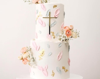 May include: A two-tiered white cake decorated with pink and blue brushstrokes, gold leaf accents, and a gold cross on top. The cake is adorned with delicate pink and white flowers.
