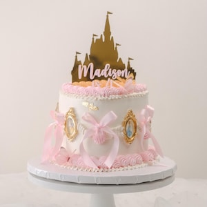 May include: A white cake decorated with pink frosting and gold accents. The cake is topped with a gold castle silhouette with the name "Madison" written in pink letters. The cake is on a white cake stand.