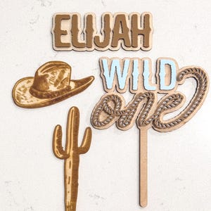 May include: Wooden cake topper set featuring a cowboy hat, cactus, and the words "Elijah Wild One" in brown and blue.