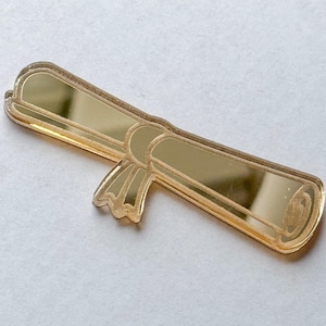 May include: A gold-colored, scroll-shaped pin with a ribbon detail. The pin has a reflective surface and a detailed outline, suggesting a graduation or achievement theme. The pin is approximately 2 inches long.