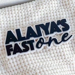 May include: Black and white plastic sign with the text "Alaiya's Fast One" in a bold, stylized font.