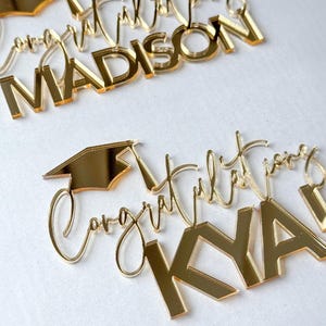 May include: Gold acrylic cake toppers with the words "Congratulations" and a name, plus a graduation cap. The toppers are in a script font and block letters. The name "Madison" is visible on one topper, and "Kya" on another.