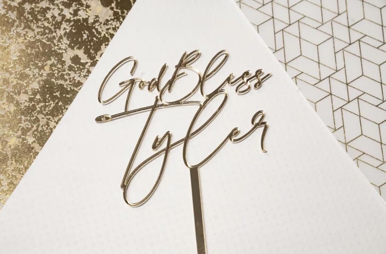 May include: Gold cake topper with the text "God Bless Tyler" in a cursive font.