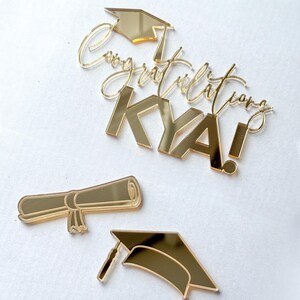 May include: A gold mirror-finish acrylic cake topper set featuring the text Congratulations KYA! with a graduation cap icon, a separate rolled diploma scroll, and a standalone graduation cap, all with a reflective metallic surface.