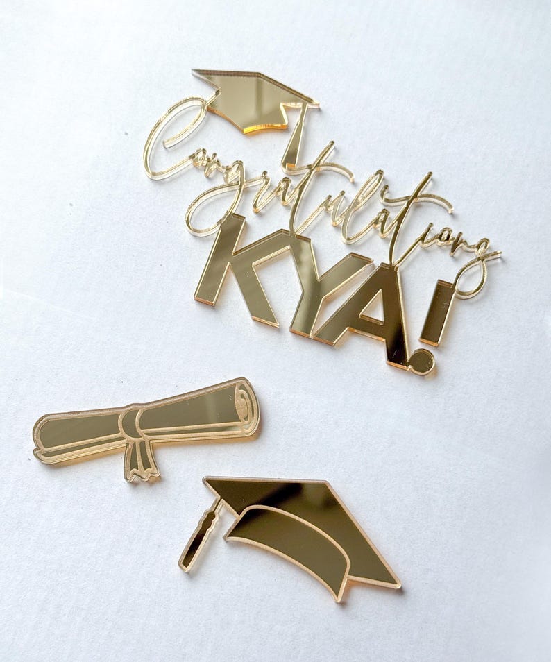 May include: A gold mirror-finish acrylic cake topper set featuring the text Congratulations KYA! with a graduation cap icon, a separate rolled diploma scroll, and a standalone graduation cap, all with a reflective metallic surface.