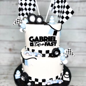 May include: Two-tiered white cake decorated with a checkered racing theme. The top tier has a black banner with the text "GABRIEL IS TWO FAST". The bottom tier has a checkered pattern and blue race cars. The cake is decorated with black and white checkered flags, blue and silver balloons, and black tire toppers.