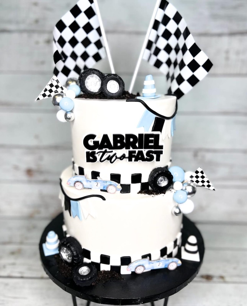 May include: Two-tiered white cake decorated with a checkered racing theme. The top tier has a black banner with the text "GABRIEL IS TWO FAST". The bottom tier has a checkered pattern and blue race cars. The cake is decorated with black and white checkered flags, blue and silver balloons, and black tire toppers.