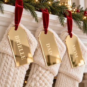 May include: Three cream-colored knit Christmas stockings hanging from a mantel, each featuring a rectangular gold mirror acrylic gift tag. The tags are suspended by red velvet ribbons and are engraved with the names BRIELLE, DAD, and MOM.