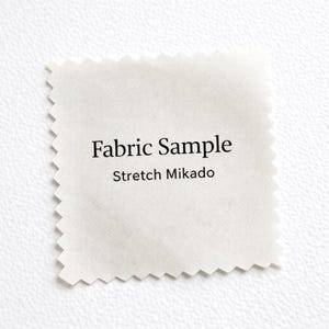 Puede incluir: Muestra de tela cuadrada con superficie blanca y texturizada. El texto "Fabric Sample" y "Stretch Mikado" está impreso en negro. Los bordes de la tela están cortados en zigzag.