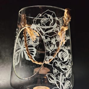 Op de afbeelding: Transparant glazen terrarium met geëtste rozen- en vlindermotieven. Een natuurlijke touwhandgreep is aan de bovenkant bevestigd. Het terrarium is ontworpen om een kleine plant of kaars te bevatten.