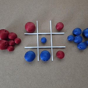 May include: A tic-tac-toe game set with red and blue game pieces. The game board is white, and the pieces are spherical with small holes. The red pieces are clustered on the left, and the blue pieces are on the right.