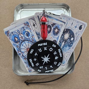 May include: A divination set featuring a black planchette with zodiac symbols and the words "Yes", "No", and "Maybe". A red crystal pendant hangs above the planchette, with a deck of illustrated cards in the background. The set is in a silver tin.