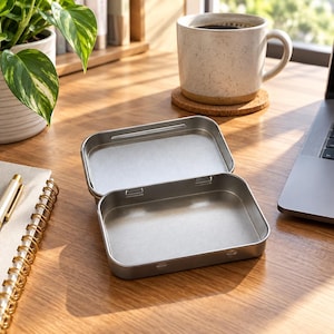 May include: A rectangular, silver-colored metal tin with a hinged lid that is open. The container has a smooth, matte finish on the interior and exterior surfaces, featuring rounded corners and a simple, functional design for storage purposes.