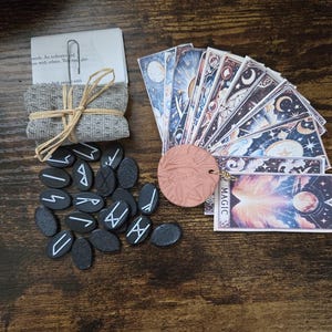 May include: An assortment of divination tools on a wooden surface. Includes a deck of tarot cards with celestial illustrations, a set of black rune stones with white symbols, a small bundle wrapped in twine, and a pink, circular charm with a floral design. A card with the word "MAGIC" is also present.