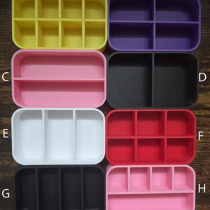 May include: A collection of eight colorful, rectangular organizers. Each organizer is divided into compartments. Colors include yellow, purple, pink, black, white, red, and two shades of pink and black. The organizers are made of a smooth material.