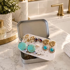 May include: A silver-toned metal storage tin containing a white foam insert with pre-cut holes. The insert holds several pairs of earrings, including large round turquoise stones, gold-toned floral designs, and small iridescent cabochon studs.