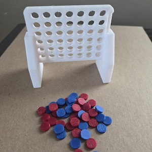 May include: A white plastic Connect Four game set. The game board has a grid of holes, and a pile of red and blue game pieces are scattered below. The game is designed for two players.