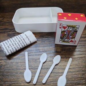 May include: A miniature game set featuring a white divided container, a deck of cards with a King of Hearts, four small white spoons, and a rolled-up paper with text. The set is arranged on a wooden surface.