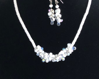 May include: A white beaded necklace with a cluster of iridescent white beads in the center. Matching earrings with iridescent white beads dangle from silver hooks.