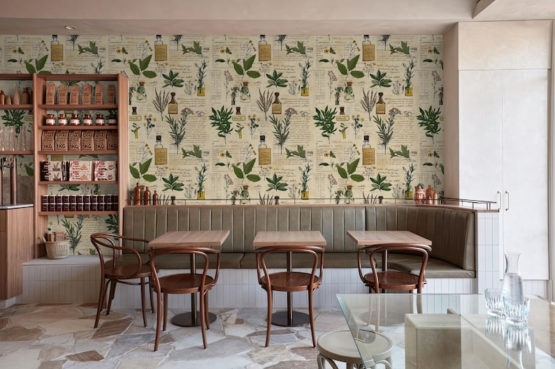May include: A restaurant interior with a botanical wallpaper featuring illustrations of plants and bottles. The wallpaper is cream-colored with green and brown accents. There are four tables with chairs in the foreground, and a wooden shelf with jars and bottles on the left side of the image.
