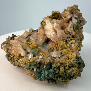 May include: A natural mineral specimen featuring a cluster of crystalline formations. The piece displays a mix of colors, including clear, beige, yellow, and green hues. The texture appears rough and uneven, showcasing the raw beauty of the stone.