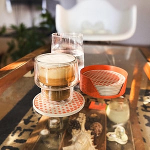 May include: A double-walled glass coffee cup filled with a layered beverage, resting on a matching coaster. A stack of coasters with a red and white pattern sits in an orange holder. A glass of water is also present.