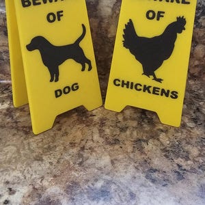 Puede incluir: Dos señales de advertencia amarillas con texto negro y siluetas. La señal de la izquierda dice "Cuidado con el perro" con una silueta de un perro. La señal de la derecha dice "Cuidado con las gallinas" con una silueta de una gallina.