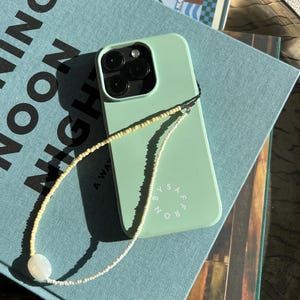 以下が含まれることがあります： 白と黄色のビーズのストラップが付いた薄い青色の携帯電話ケース。ケースには「Around」と「Safe」の文字が印刷されています。携帯電話ケースは、「Morning」という文字が印刷された青い本のの上に置かれています。