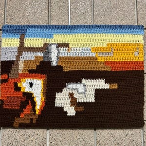 May include: Crocheted wall hanging depicting a famous painting. The artwork features a brown, orange, and yellow sunset with two white figures in the foreground.