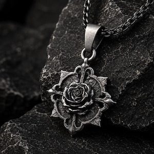 May include: A dark silver-toned metal pendant necklace featuring a detailed three-dimensional rose at the center of an ornate, star-shaped frame with pointed edges. The pendant hangs from a matching dark silver-toned chain link necklace.