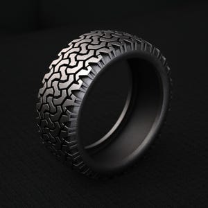 May include: A dark gray metal ring featuring a textured outer surface that mimics the tread pattern of an off-road tire. The band has a wide, flat profile with deep, repeating geometric grooves carved into the exterior, creating a rugged appearance.